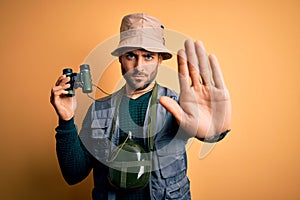 Young handsome tourist man with beard on vacation wearing explorer hat using binoculars with open hand doing stop sign with