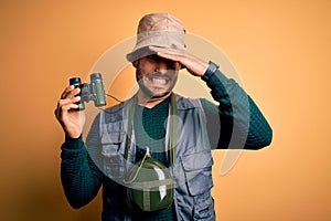 Young handsome tourist man with beard on vacation wearing explorer hat using binoculars stressed with hand on head, shocked with