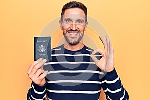 Young handsome tourist man holding united states passport over isolated yellow background doing ok sign with fingers, smiling