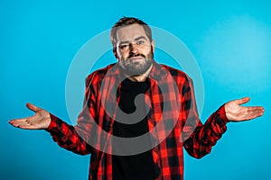 Young handsome unsure guy in red plaid shirt shrugs his arms, makes gesture of I don`t know, can`t help anything