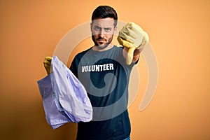 Young handsome volunteer man with beard cleaning junk using bag over yellow background with angry face, negative sign showing