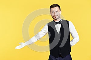 Young handsome waiter doing a welcome gesture over yellow background