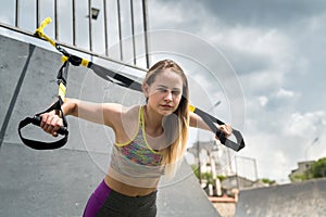 Young handsome woman in sportswear doing push ups training arms with trx fitness straps at nature