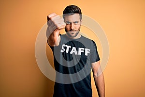 Young handsome worker man with beard wearing staff uniform t-shirt over yellow background with angry face, negative sign showing