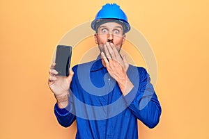 Young handsome worker man wearing uniform and hardhat holding smartphone showing screen covering mouth with hand, shocked and