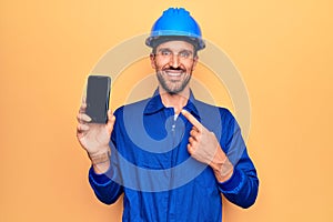 Young handsome worker man wearing uniform and hardhat holding smartphone showing screen smiling happy pointing with hand and