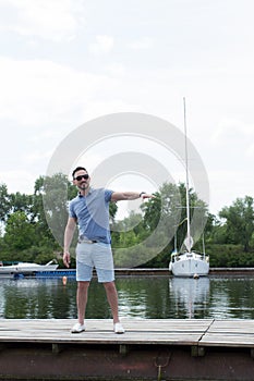 Young handsome yachtsman in sun glasses walking at river pier and welcomes friends. Handsome businessman invites to his boat.