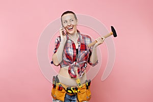 Young handyman woman in plaid shirt, kit tools belt full of instruments talking on mobile phone, conducting pleasant