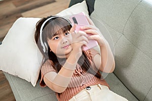 Young happy and adorable Asian girl is using a smartphone while lying on a sofa in the living room