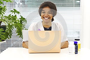 Young happy African-American female doctor sitting at workplace looking to the camera