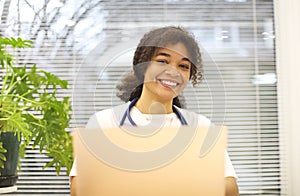 Young happy African-American female doctor sitting at workplace looking to the camera