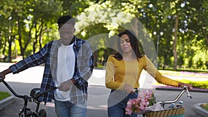 Young happy african american man and woman walking with bicycles, laughing together in urban park, tracking shot