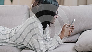 Young happy african american woman lying on sofa relaxing at home rests chatting online using phone makes purchase on