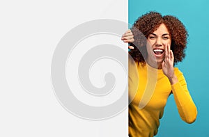 Young happy African American woman peeking out from empty white board and holding her hands near her mouth scream