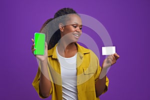 Young happy African American woman shows phone and credit card stands in studio