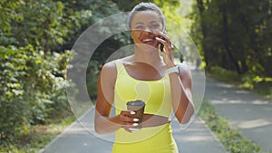 Young happy african american woman walking in morning park, carrying takeaway coffee and talking on cellphone