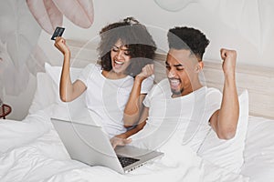 Young happy african loving couple girl guy in white t-shirts using laptop pc credit bank card lying on bed in bedroom at