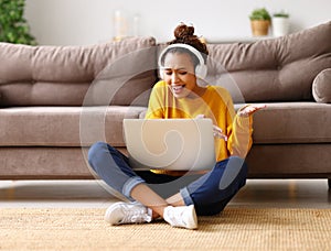 Happy african american teen girl in headphones sitting on floor and  having video call while working or studying online on sofa at