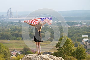 Young happy american woman with long hair raising up waving on wind USA national flag in her hands relaxing outdoors enjoying warm