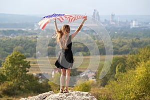 Young happy american woman with long hair raising up waving on wind USA national flag in her hands relaxing outdoors enjoying warm