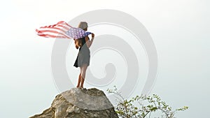 Young happy american woman with long hair raising up waving on wind USA national flag in her hands relaxing outdoors enjoying warm