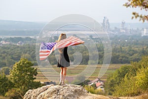 Young happy american woman with long hair raising up waving on wind USA national flag in her hands relaxing outdoors enjoying warm