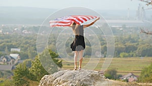 Young happy american woman with long hair raising up waving on wind USA national flag in her hands relaxing outdoors enjoying warm