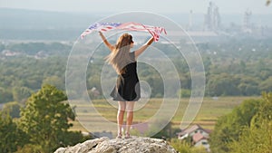Young happy american woman with long hair raising up waving on wind USA national flag in her hands relaxing outdoors enjoying warm