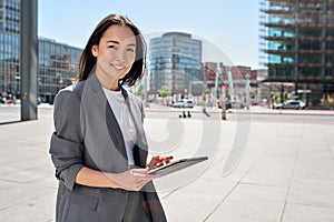 Young happy Asian business woman standing on city street holding digital tablet.