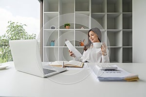 Young happy asian business woman working at office computer desk smiling drinking cup of coffee relaxed