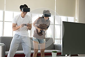 Young happy Asian couple playing video games wearing virtual reality glasses in their apartment