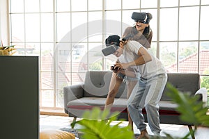 Young happy Asian couple playing video games wearing virtual reality glasses in their apartment