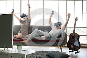 Young happy Asian couple playing video games wearing virtual reality glasses in their apartment