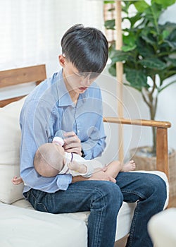 Young happy asian father holding and feeding newborn baby with milk bottle sitting on sofa at home or apartment. Smiling and