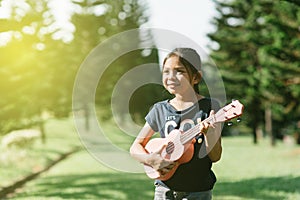 Young and happy asian girl playing ukelele guitar in the park at sunny morning while looking to copy space