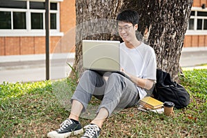 Young happy Asian male university student is using his laptop under the tree in a campus park