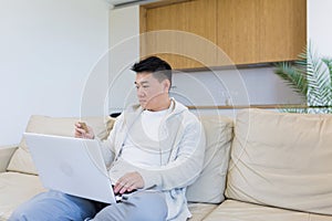Young happy asian man shopping online in internet store using laptop computer and credit card. male enters data of paying for
