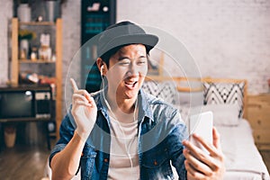 Young happy Asian man talking video call via smartphone wearing headphones at home.