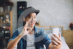 Young happy Asian man talking video call via smartphone wearing headphones at home.