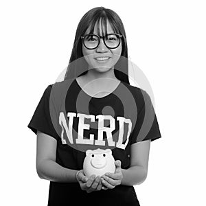 Young happy Asian nerd teenage girl smiling and holding piggy bank