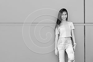 Young happy Asian teenage girl smiling while leaning against concrete wall outdoors