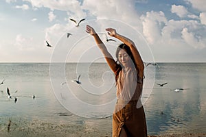 Happy asian woman with hands in the air walks on the seaside in autumn. Seagulls flying on the beach.