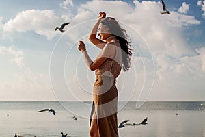 Happy asian woman with hands in the air walks on the seaside in autumn. Seagulls flying on the beach.