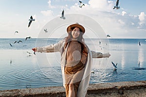 Happy asian woman with hands in the air walks on the seaside in autumn. Seagulls flying on the beach.