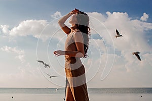 Happy asian woman with hands in the air walks on the seaside in autumn. Seagulls flying on the beach.