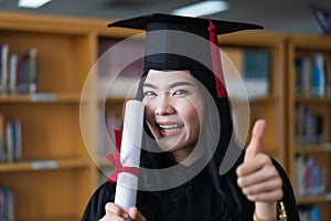 A young happy Asian woman university graduates in graduation gown and cap wears a face mask holds a degree certificate to