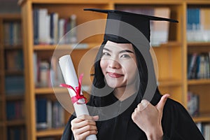 A young happy Asian woman university graduates in graduation gown and cap wears a face mask holds a degree certificate to