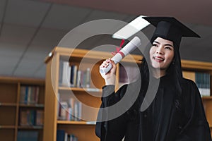 A young happy Asian woman university graduates in graduation gown and cap wears a face mask holds a degree certificate to