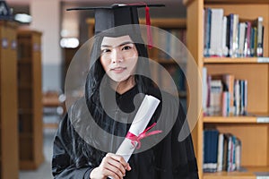 A young happy Asian woman university graduates in graduation gown and cap wears a face mask holds a degree certificate to