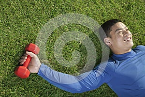 Young happy athletic man lying down in grass with red dumbbell, view from above